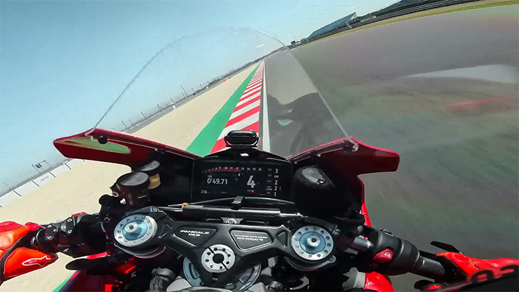 A first-person POV shot from a motorcycle leaning into a corner on a paved race track.
