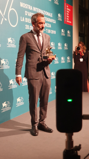 A man holding a golden lion award on stage at the 2019 Venice Film Festival, with an Insta360 ONE X recording in the foreground.
