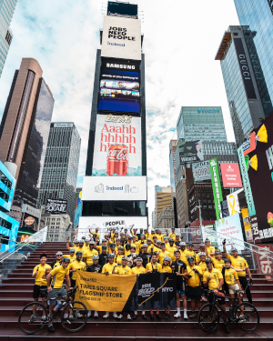 A crowd in matching Insta360 yellow shirts gathers on the red steps of Times Square holding a banner, with cyclists in front, during the flagship store launch celebration.
