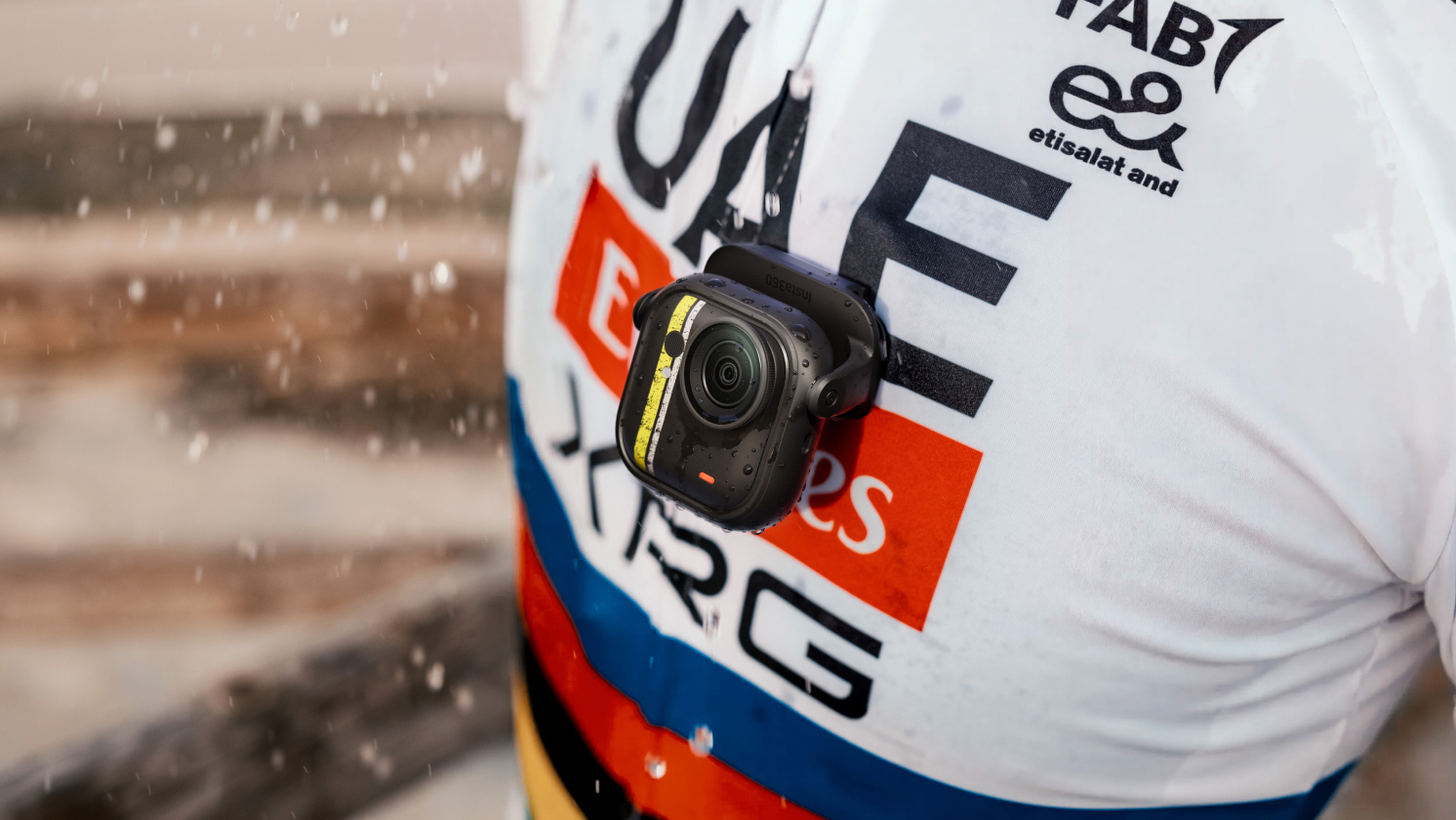 Close-up of Insta360 GO Ultra camera mounted on a cyclist’s chest, capturing raindrops on the lens and jersey during a wet ride. The rider wears the iconic white UAE Team Emirates XRG jersey.