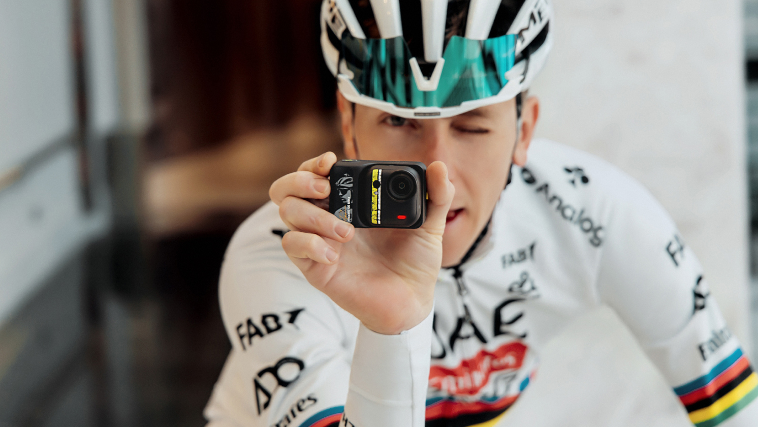 Cyclist Tadej Pogačar holding the Insta360 GO Ultra camera close to his face while wearing his white UAE Team Emirates XRG world champion jersey and helmet.